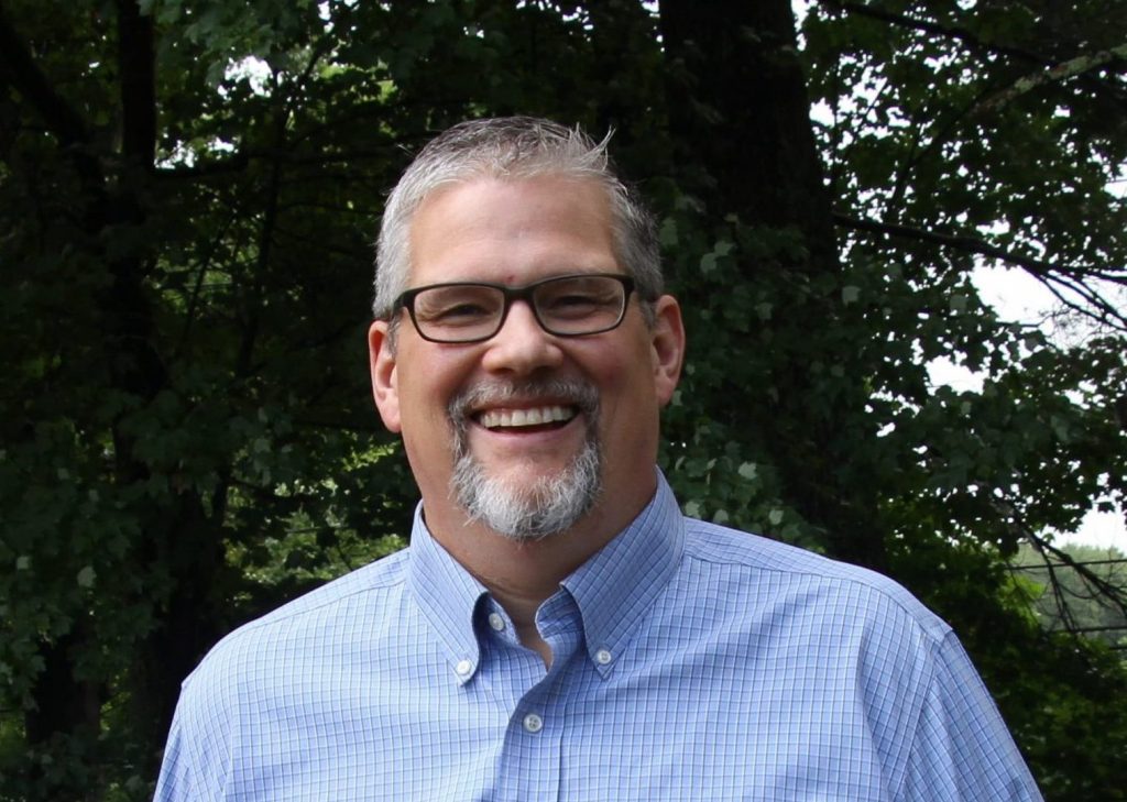 Portrait of a man wearing a blue button up shirt and black rimmed glasses with trees behind him. Bill Labich. 