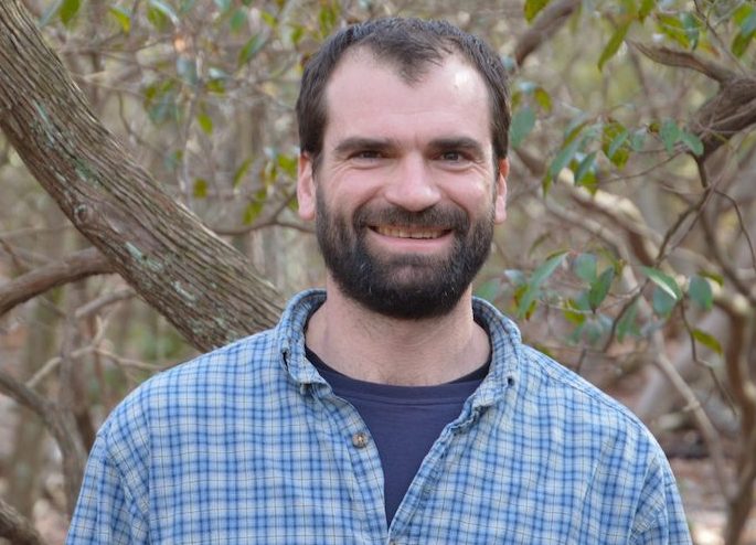 Portrait of a man wearing a blue checker button up shirt with trees behind him. Jesse Hubbard. 