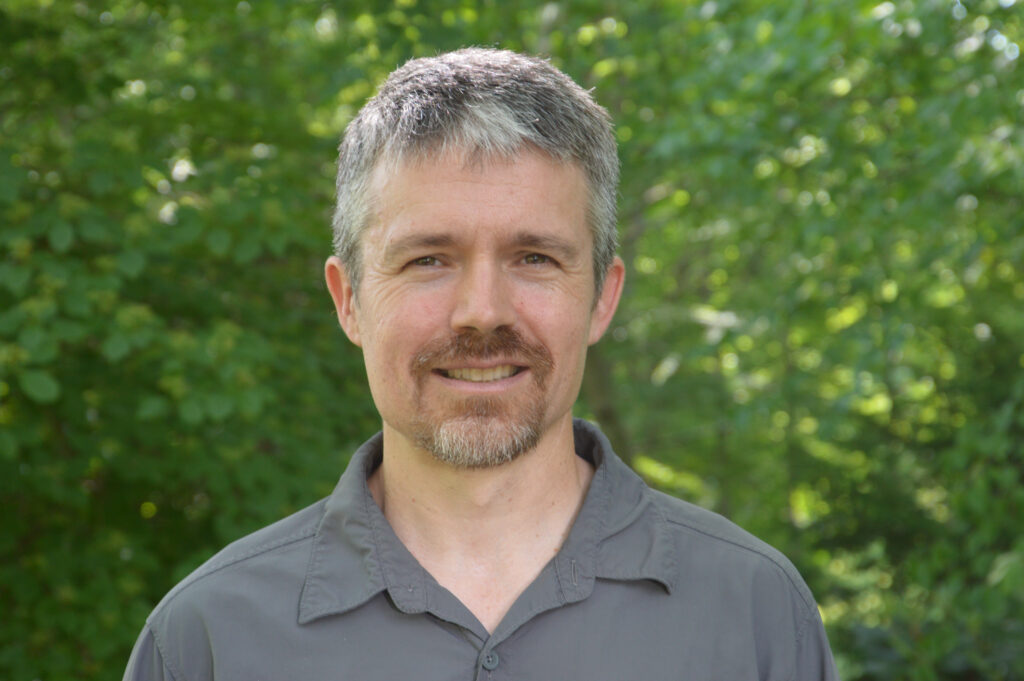 Portrait of a man wearing a grey polo shirt with trees behind him. Ed Faison. 