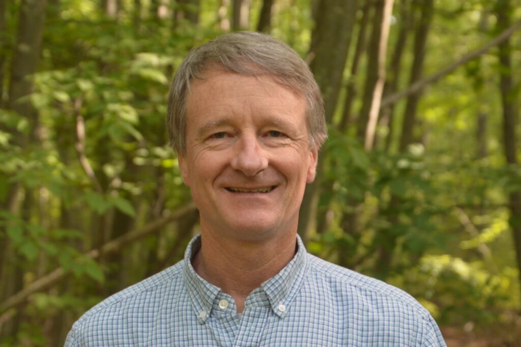 Portrait of a man and a light blue checker button up shirt with trees behind him. Geordie Elkins.  Staff Interview