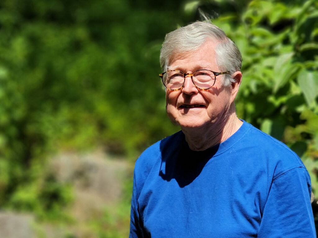 A man with grey hair, a blue shirt, and round eyeglasses outdoors.
